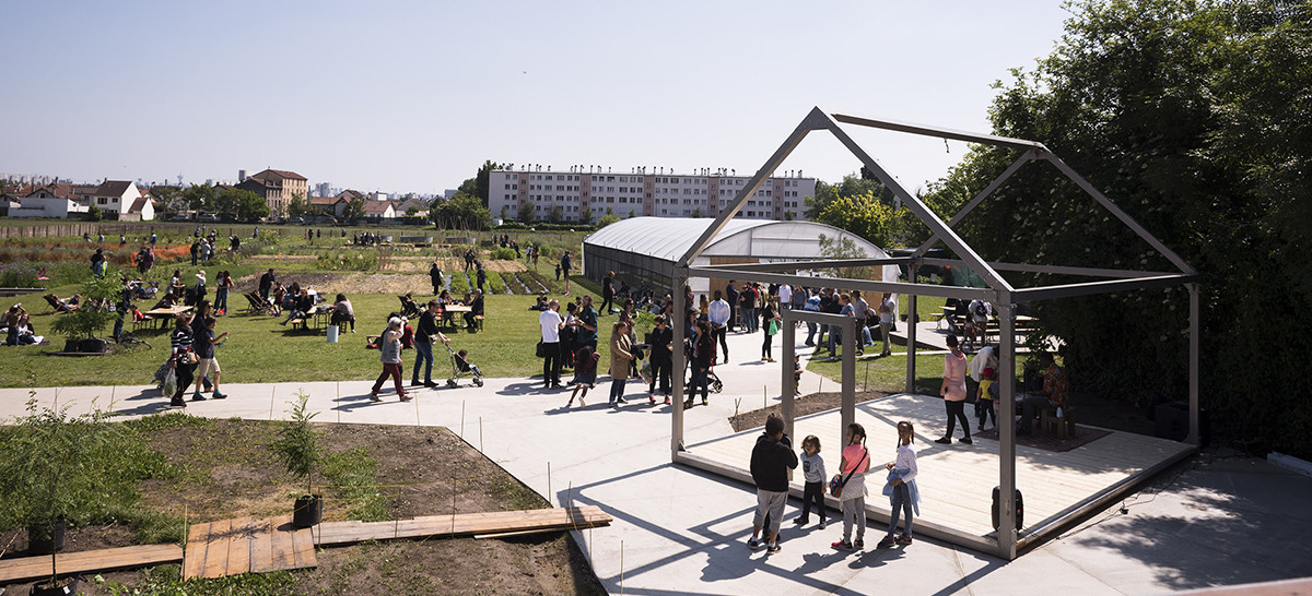 Photo montrant une journée portes ouvertes à La Ferme urbaine de Saint-Denis avec de nombreux habitants réunis pour l’occasion. On y voit de nombreuses parcelles cultivées, une serre et des immeubles d’habitation ainsi que des maisons en arrière-plan.  