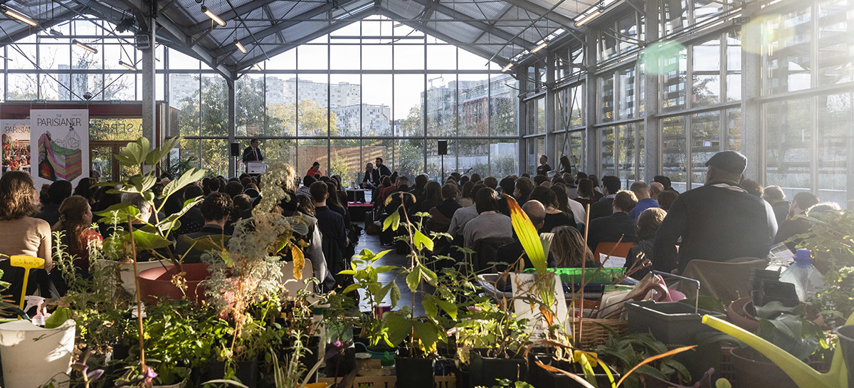 Un très grand nombre de personnes réunies à la Serre Wangari pour assister au lancement du projet Alimentaire territorial de Plaine Commune. Sur la photo les invités sont assis et assistent à une présentation qui se déroule sur une scène face à eux. Le tout a lieu dans une grande serre qui abrite des plantes.  