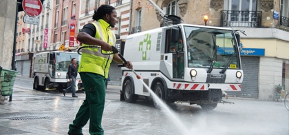 Photo montrant un agent de propreté nettoyant une rue avec deux véhicules de Plaine Commune en arrière-plan.     